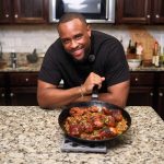 Man smiling at camera with a pan of cooked food on a kitchen counter, dark wood cabinets, microwave in background.