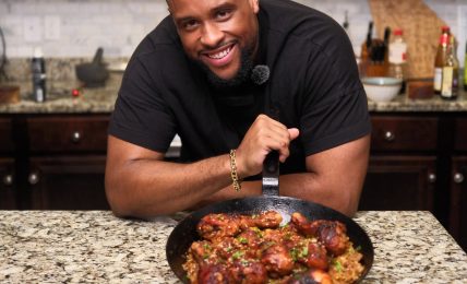 Man smiling at camera with a pan of cooked food on a kitchen counter, dark wood cabinets, microwave in background.