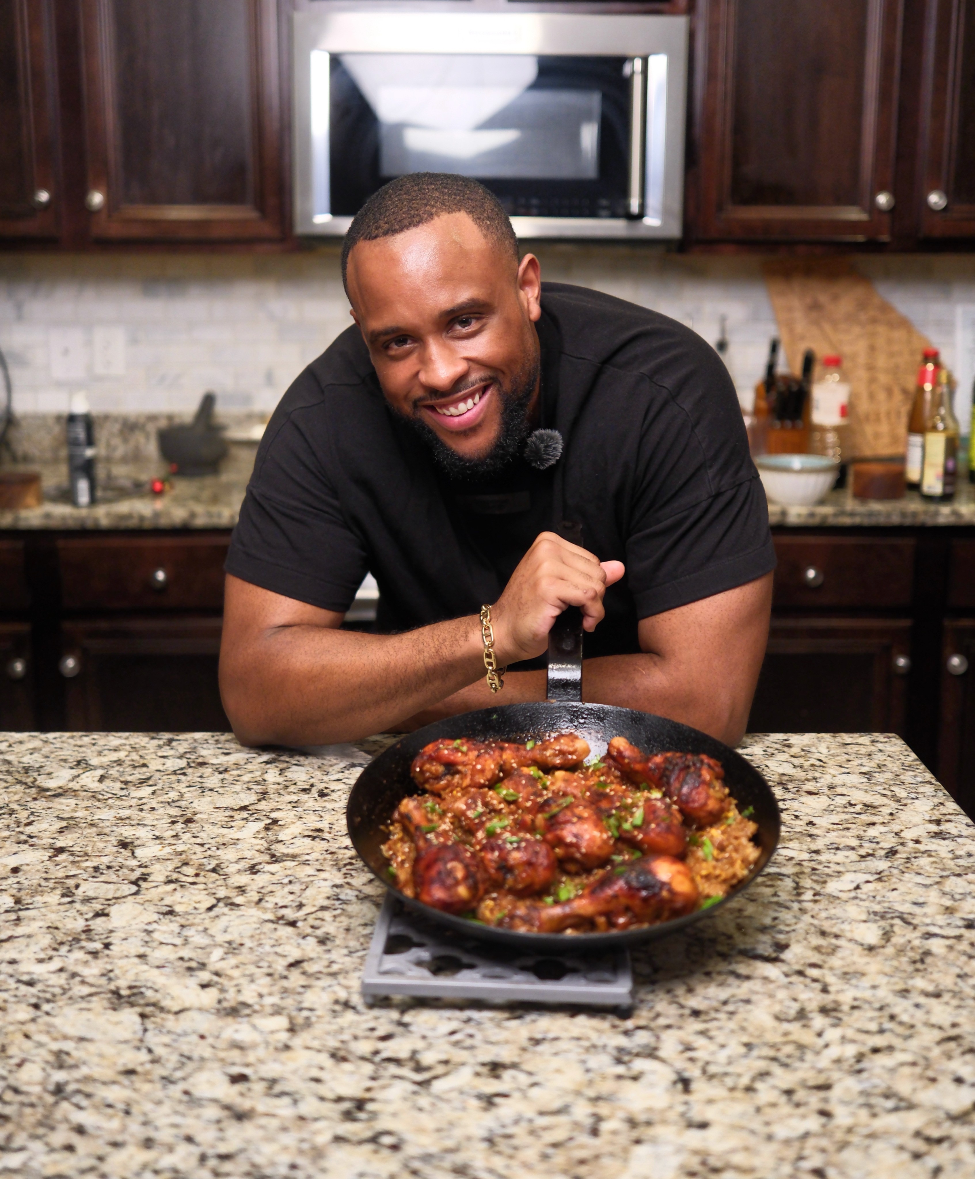 Man smiling at camera with a pan of cooked food on a kitchen counter, dark wood cabinets, microwave in background.