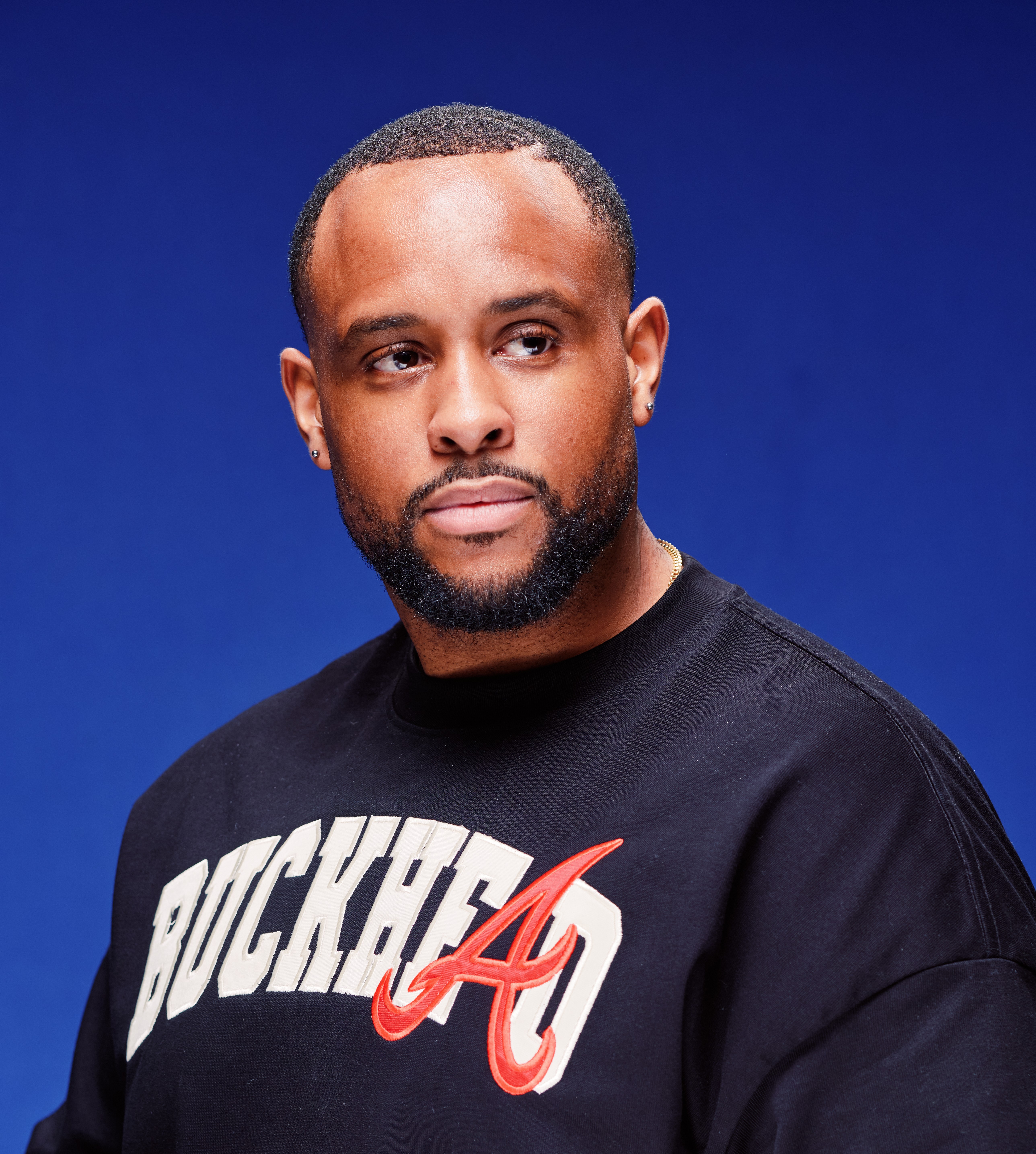 Portrait of a man with a beard and short hair wearing a black shirt with white and red text, against a blue background.