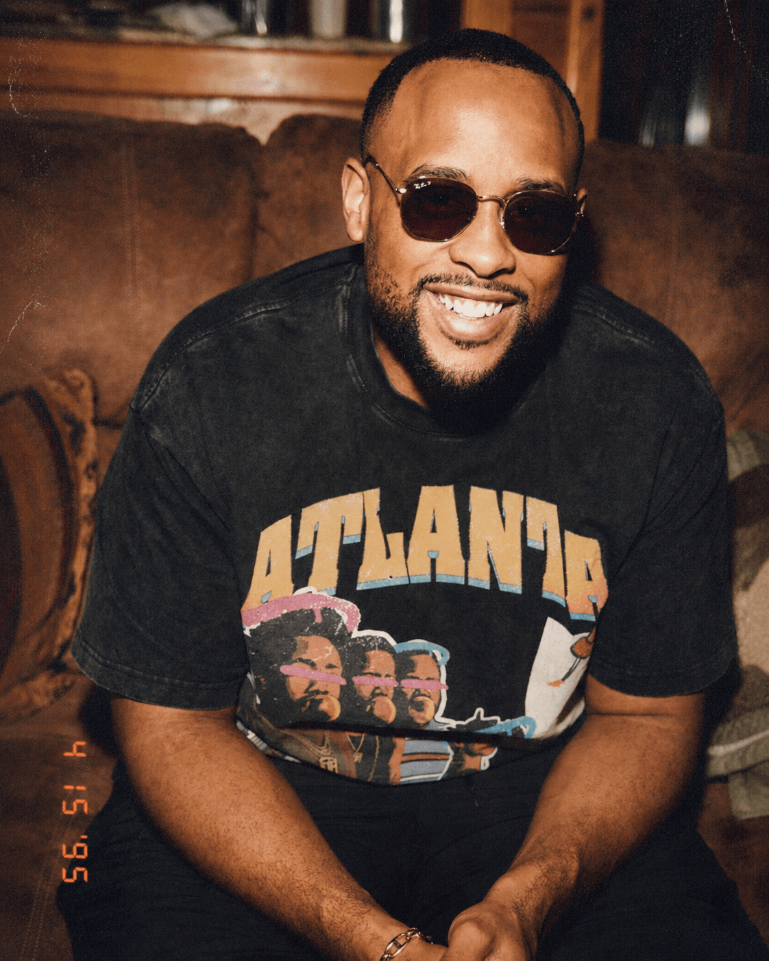 Man with sunglasses smiling, wearing a black T-shirt with 'ATLANTA' and a graphic, sitting on a brown couch.