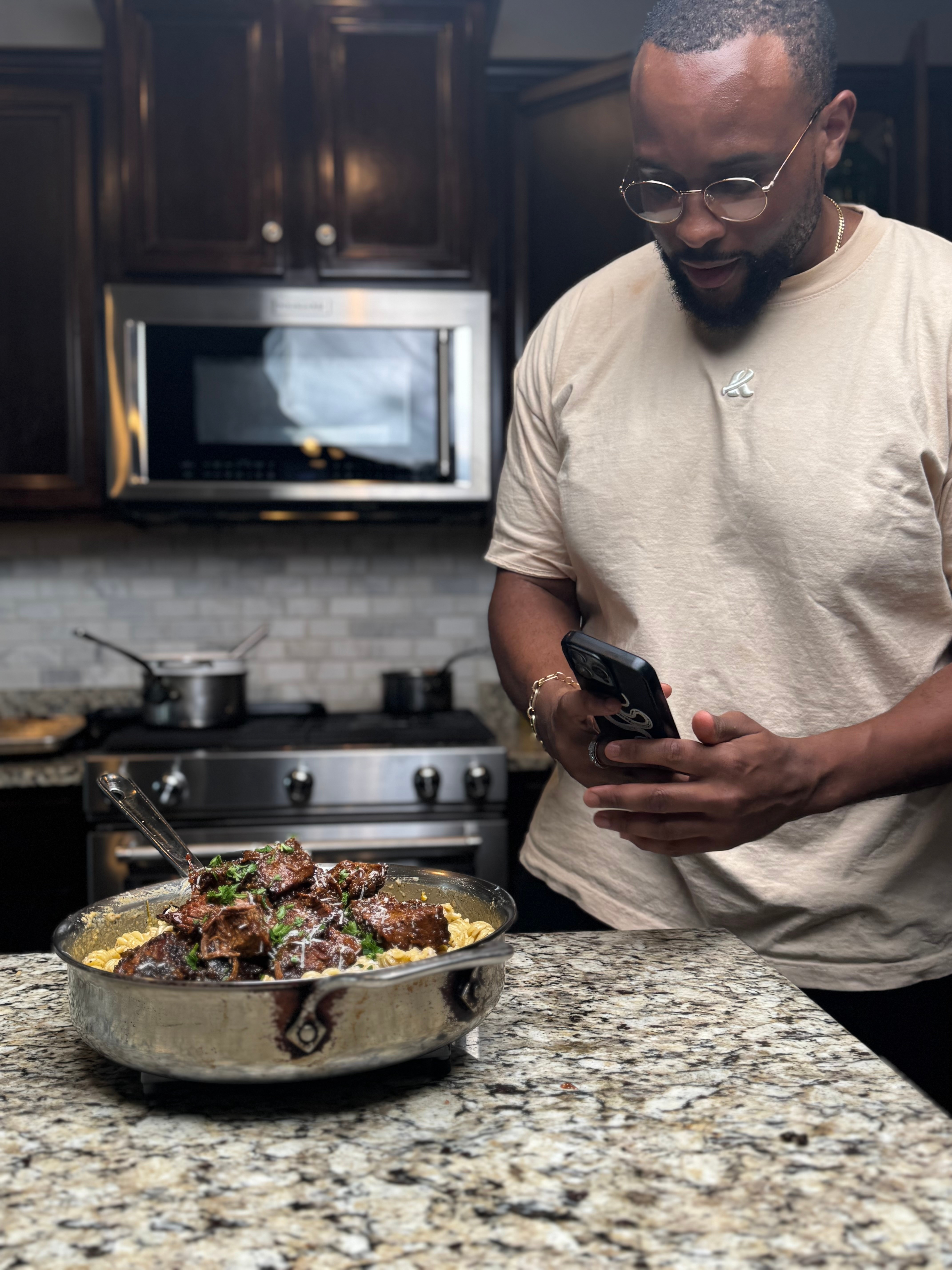 Man in glasses and beige shirt looking at phone in kitchen with stove and microwave, pot on stove, bowl of cooked food on counter