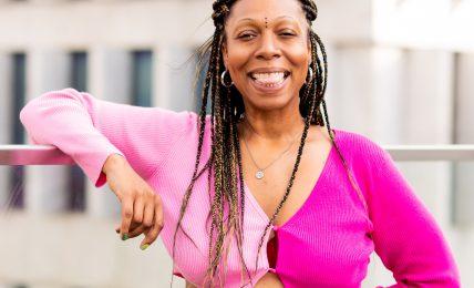 Smiling woman with long braided hair wearing a pink top and patterned skirt outdoors, with a modern building in background.