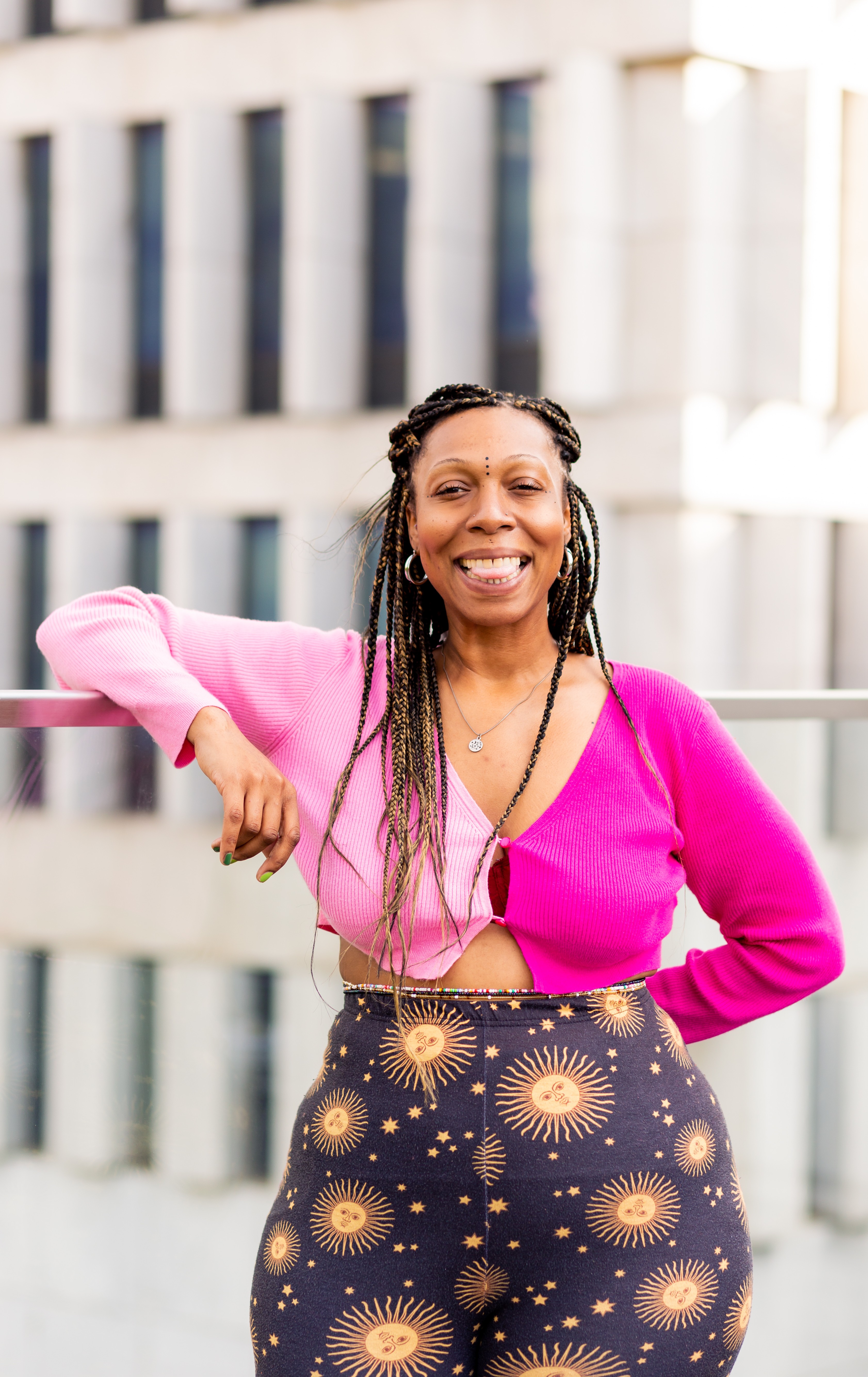 Smiling woman with long braided hair wearing a pink top and patterned skirt outdoors, with a modern building in background.