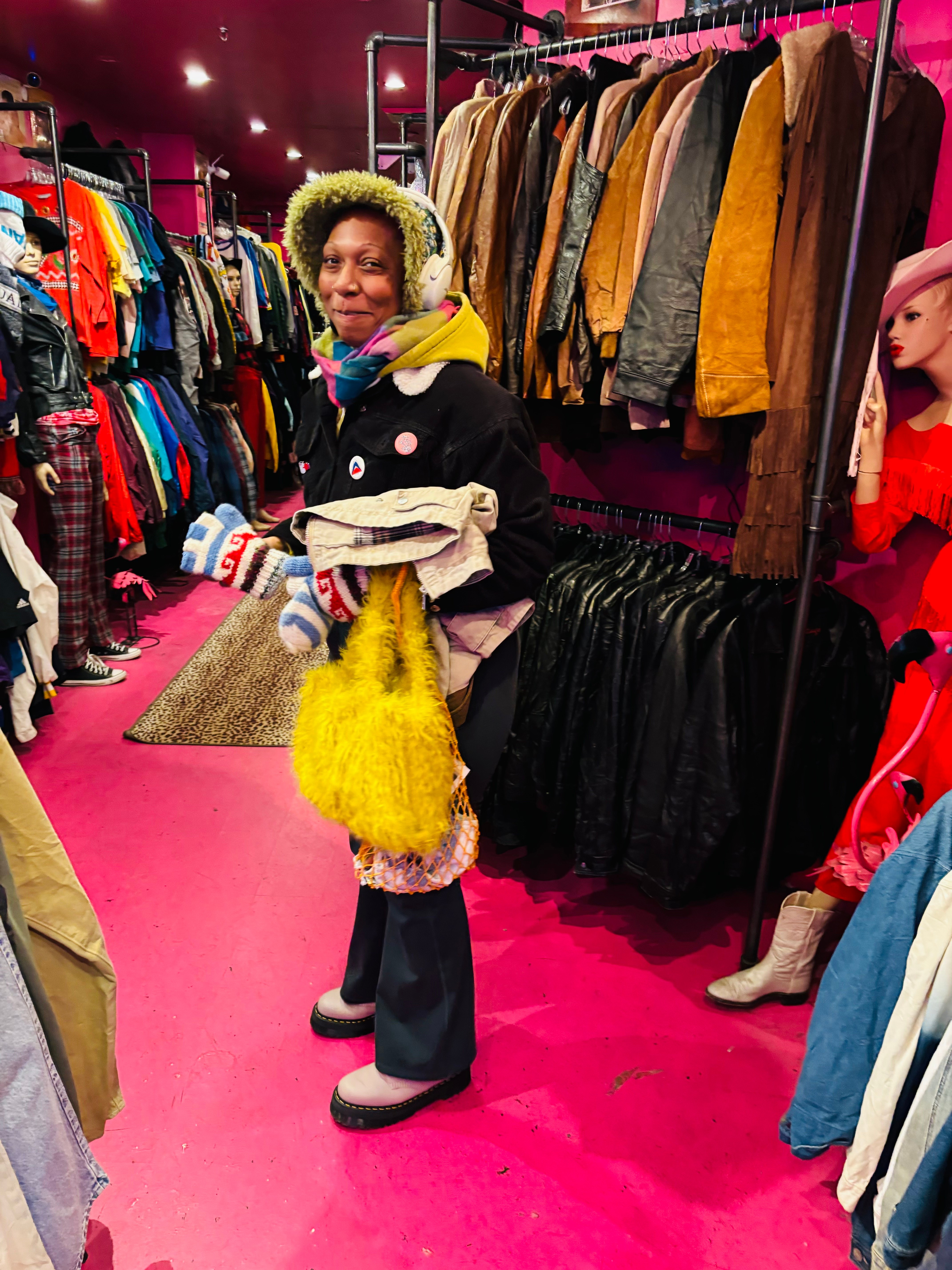Person shopping in clothing store, holding folded clothes, surrounded by racks of jackets and garments, pink floor, smiling face.