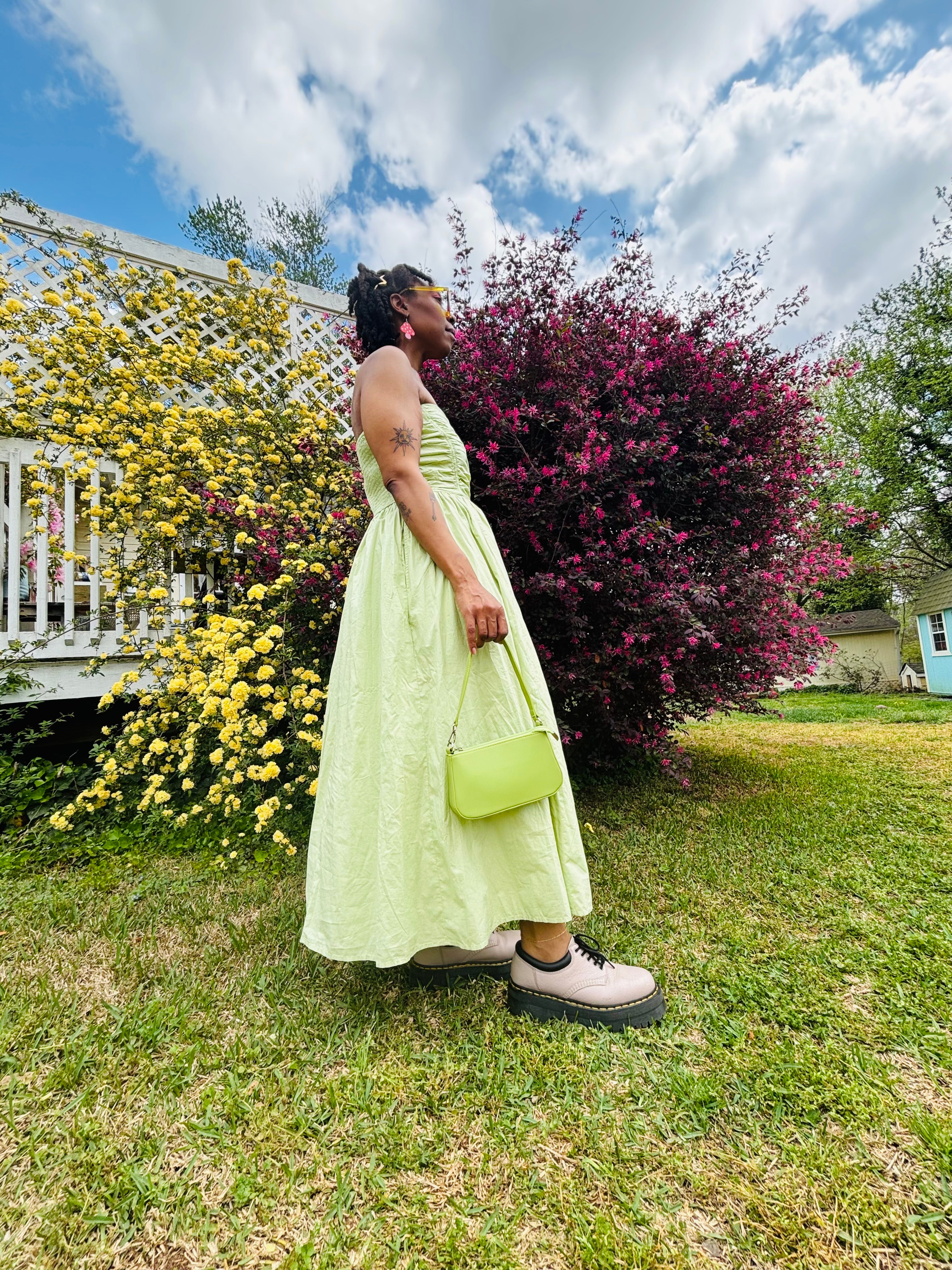 Person in a light green dress holding a matching bag, standing outdoors near colorful bushes and trees under a partly cloudy sky.