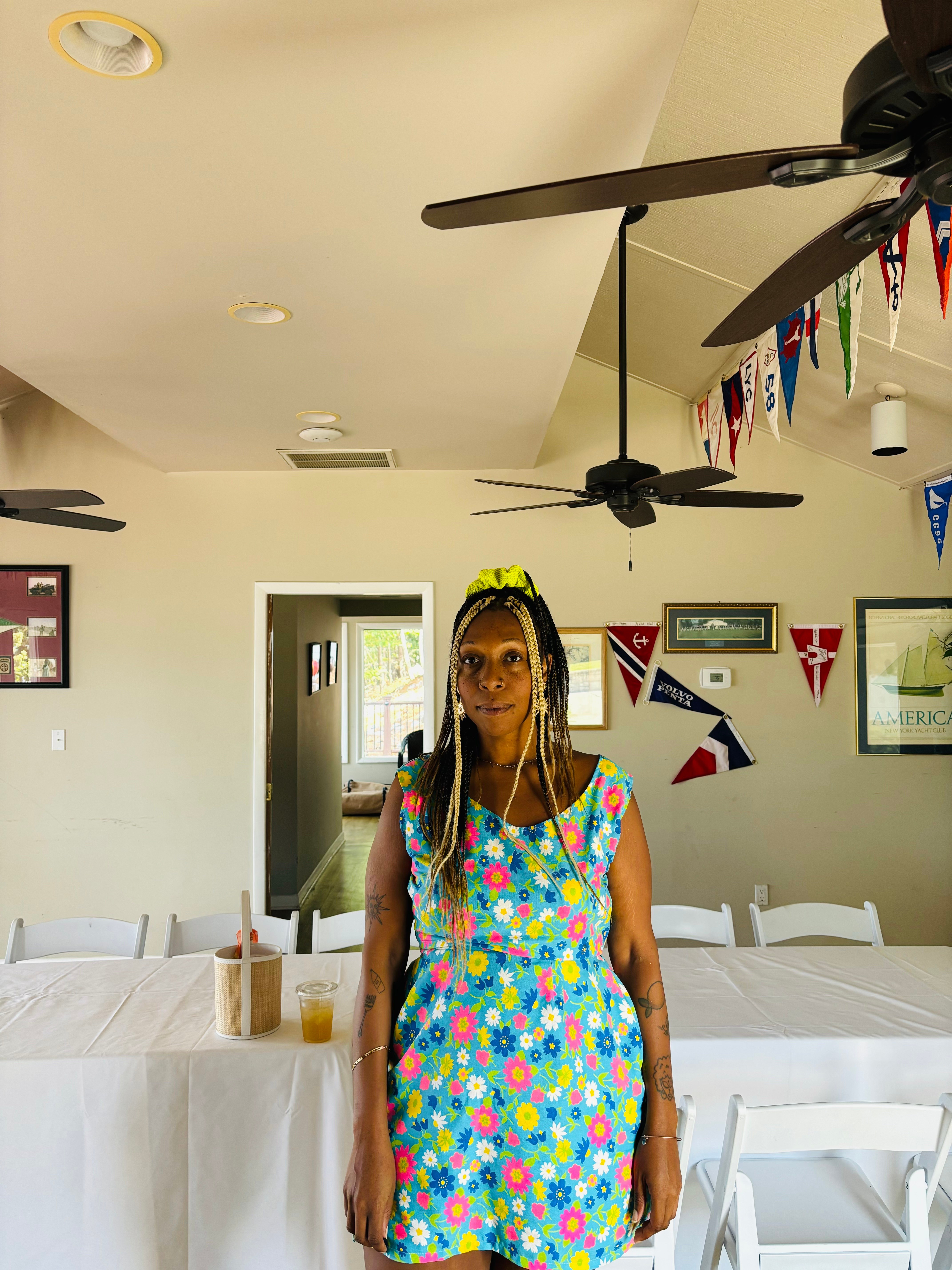Woman with long braided hair and glasses wearing a colorful floral dress standing indoors with bunting decorations on the wall.