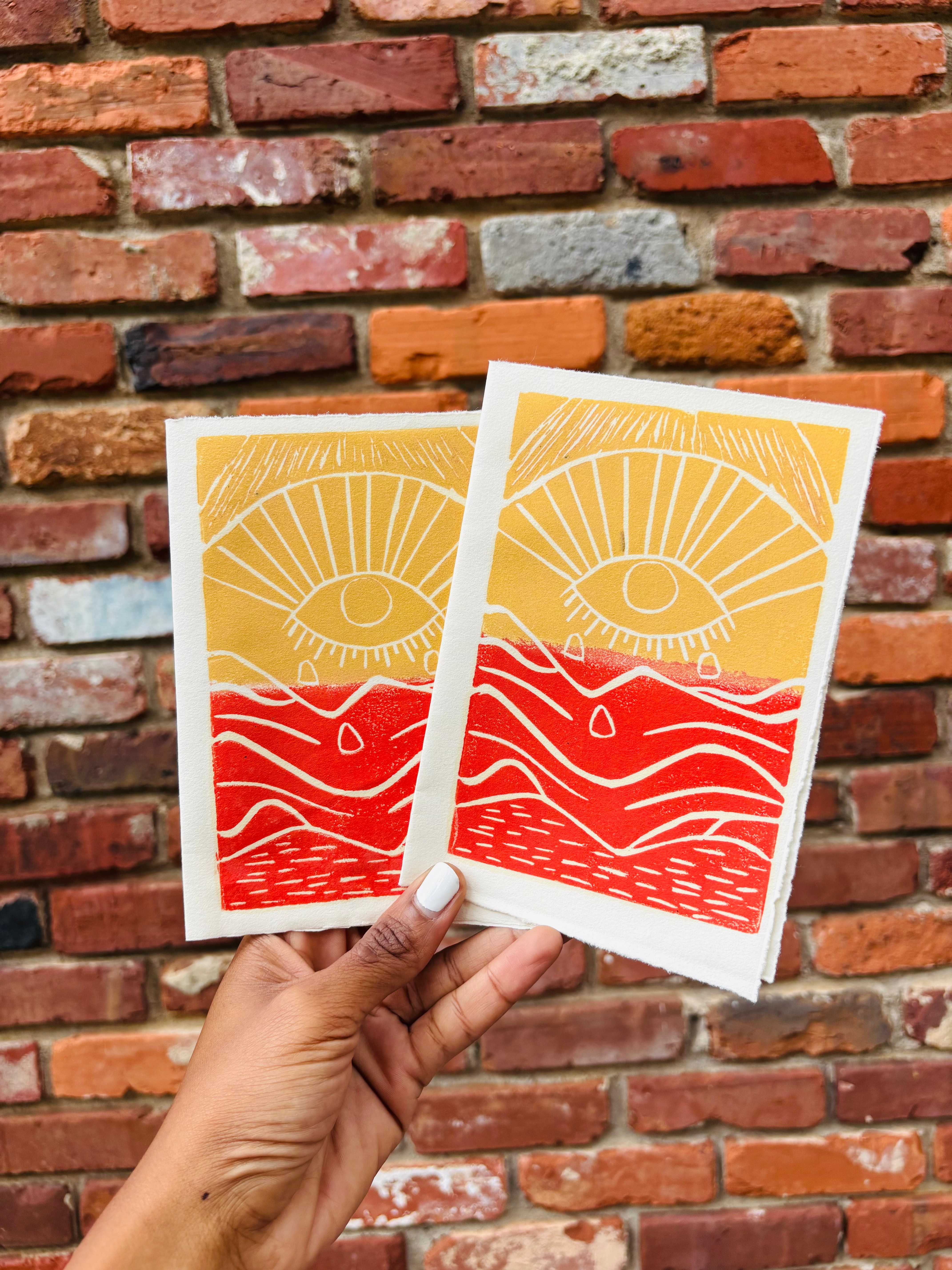Two colorful postcards with eye and wave designs held against a brick wall background.