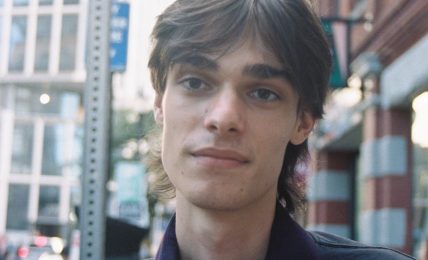 Young man with dark hair and a dark jacket stands outdoors in an urban setting, with buildings and a blue arrow sign behind him.