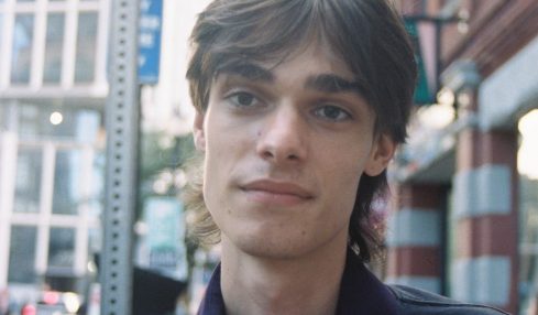 Young man with dark hair and a dark jacket stands outdoors in an urban setting, with buildings and a blue arrow sign behind him.