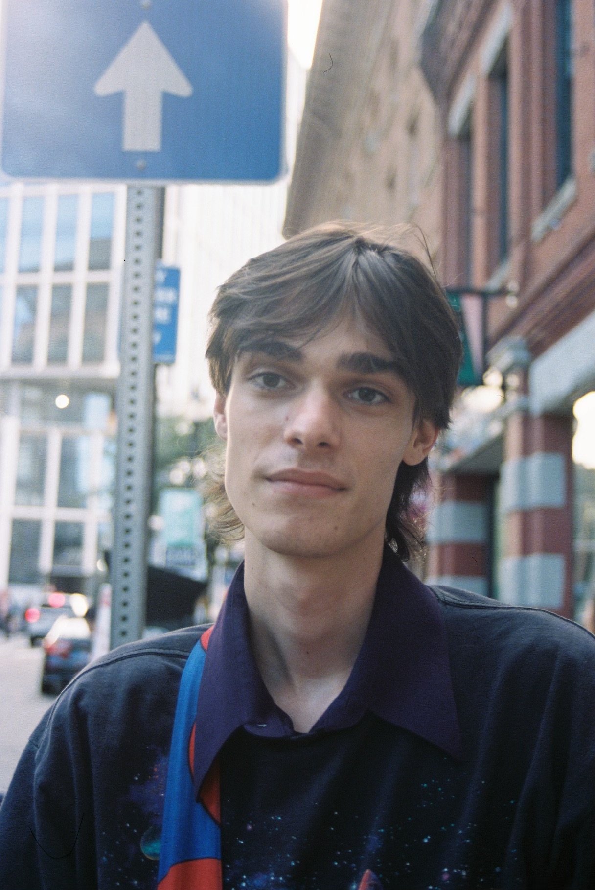 Young man with dark hair and a dark jacket stands outdoors in an urban setting, with buildings and a blue arrow sign behind him.