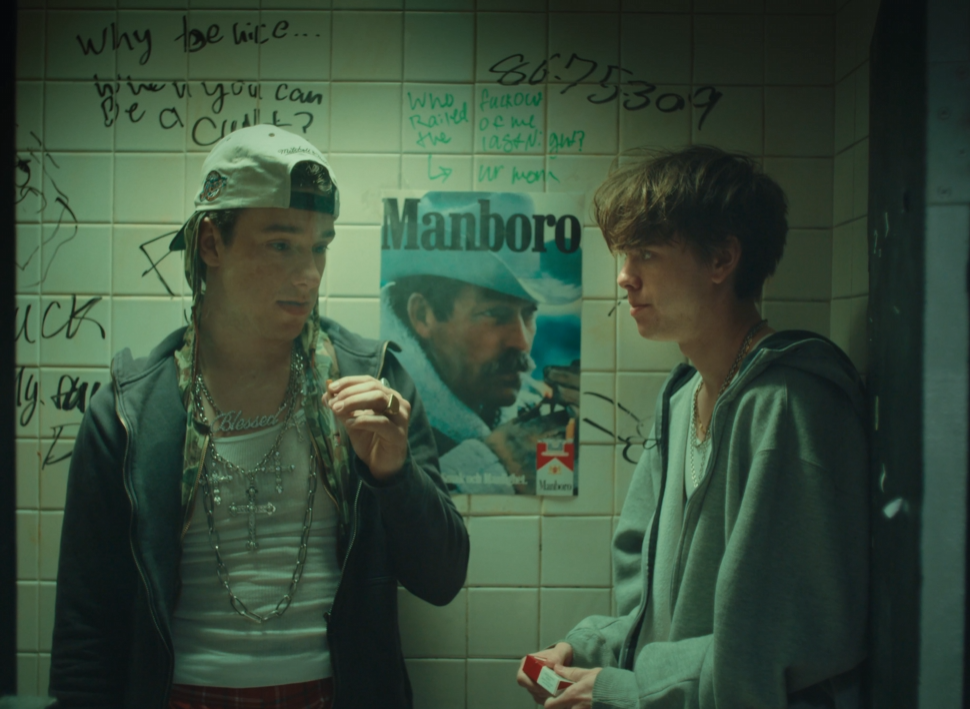 Two young people stand in front of a wall with handwritten notes and a Marlboro poster, engaging in conversation.