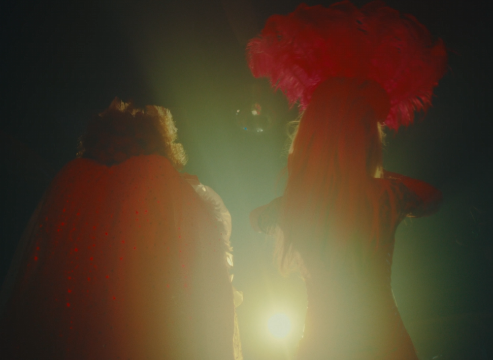 Two people with colorful wigs, one with a red wig, standing outdoors with sunlight behind them.