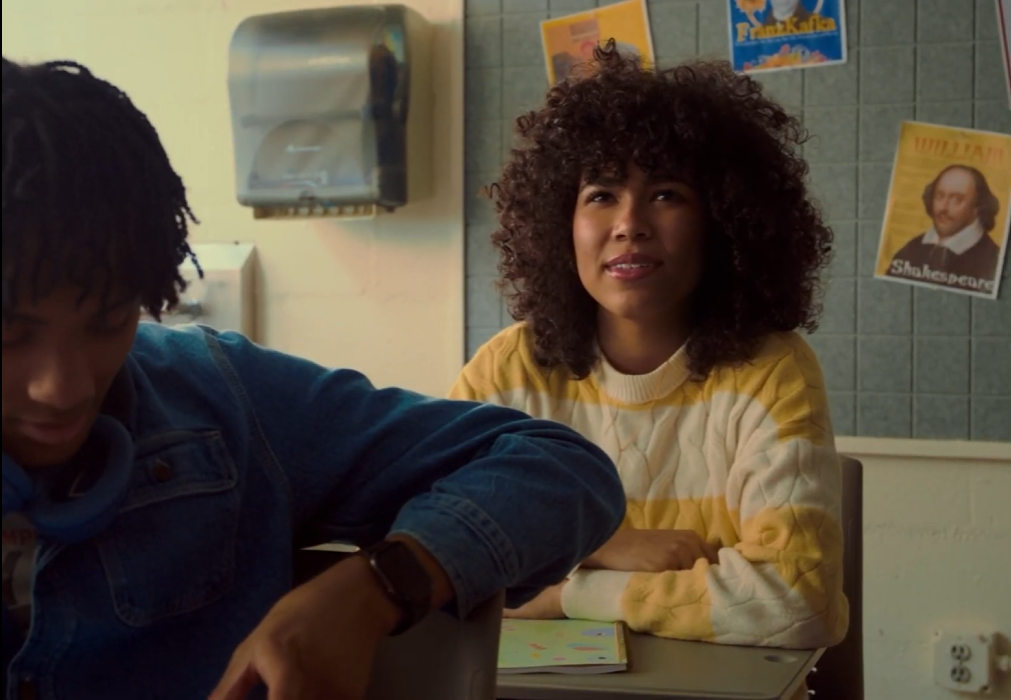 A woman with curly hair sits at a desk in a classroom, smiling, with a bulletin board behind her.
