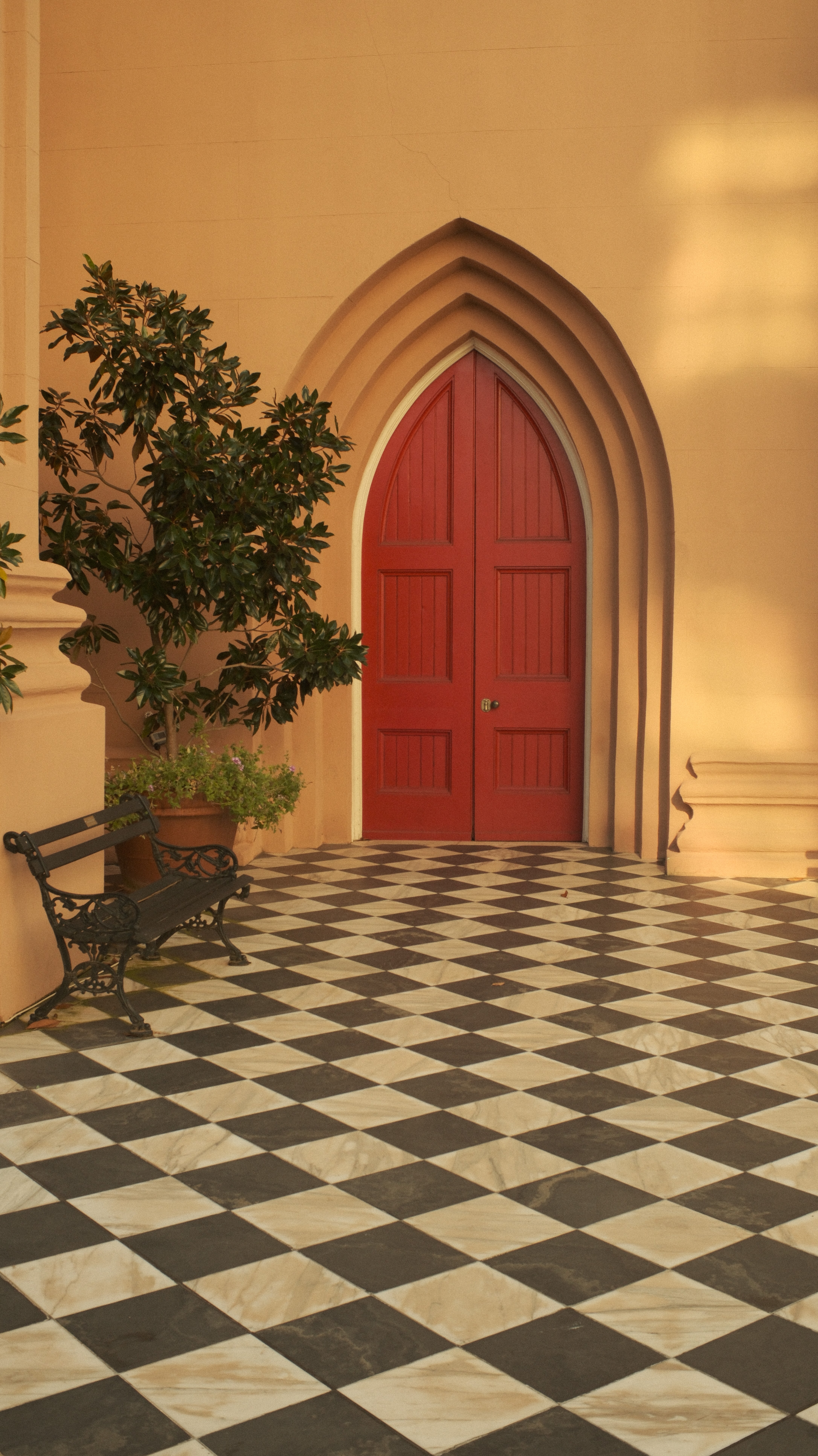 Red arched door with layered frame, potted plant on a bench, checkered floor, and warm lighting.
