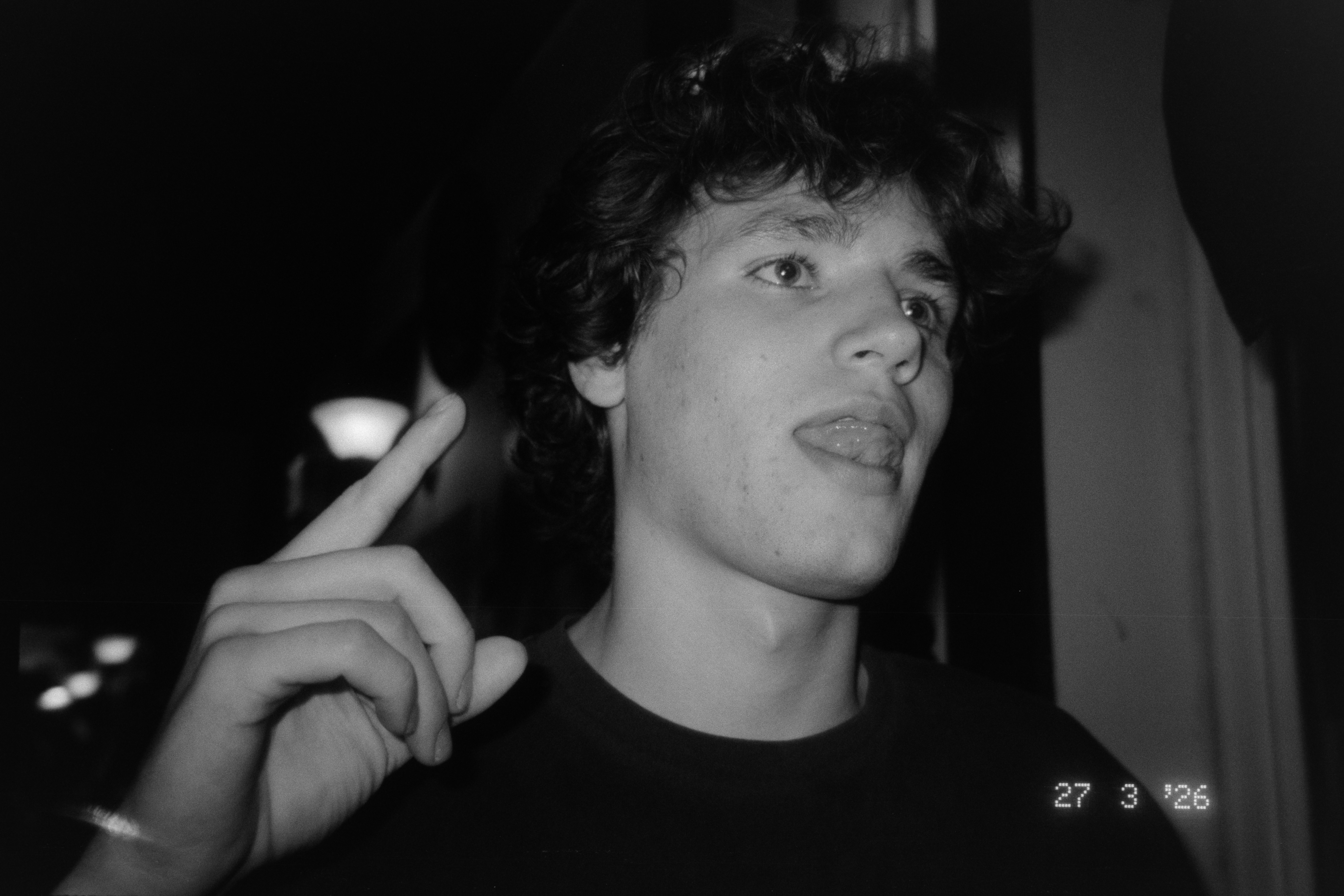 Young person with curly hair sticking out tongue and raising index finger, in black and white, indoors.
