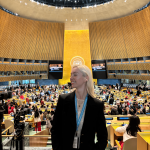 Woman with blonde hair in a black blazer stands in a large, modern conference hall with a high ceiling and many attendees.