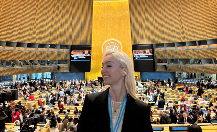 Woman with blonde hair in a black blazer stands in a large, modern conference hall with a high ceiling and many attendees.