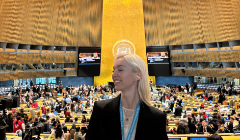 Woman with blonde hair in a black blazer stands in a large, modern conference hall with a high ceiling and many attendees.