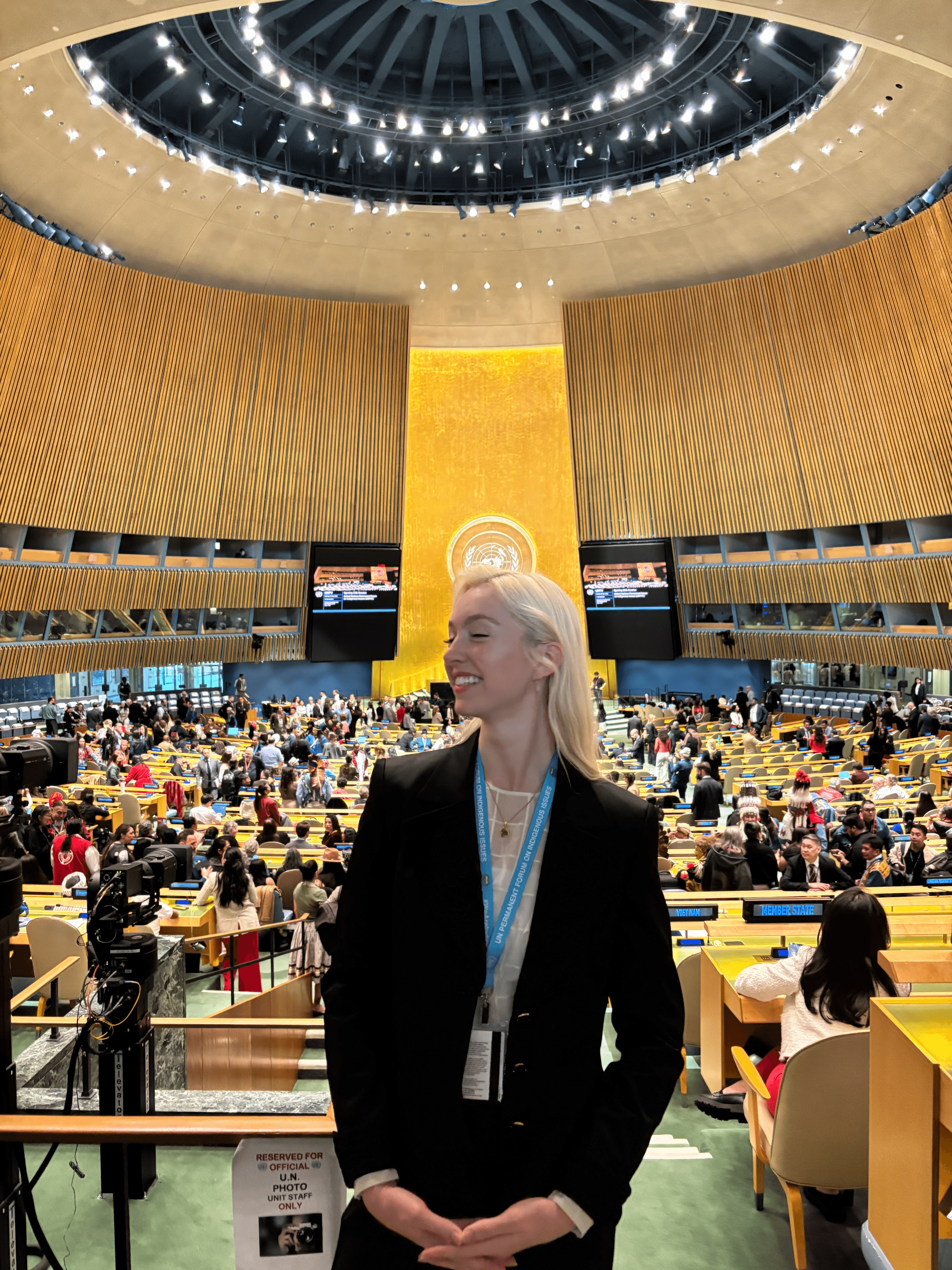 Woman with blonde hair in a black blazer stands in a large, modern conference hall with a high ceiling and many attendees.