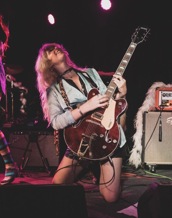 Female musician playing guitar on stage with equipment and lighting, kneeling with one knee on the ground, hair covering face.