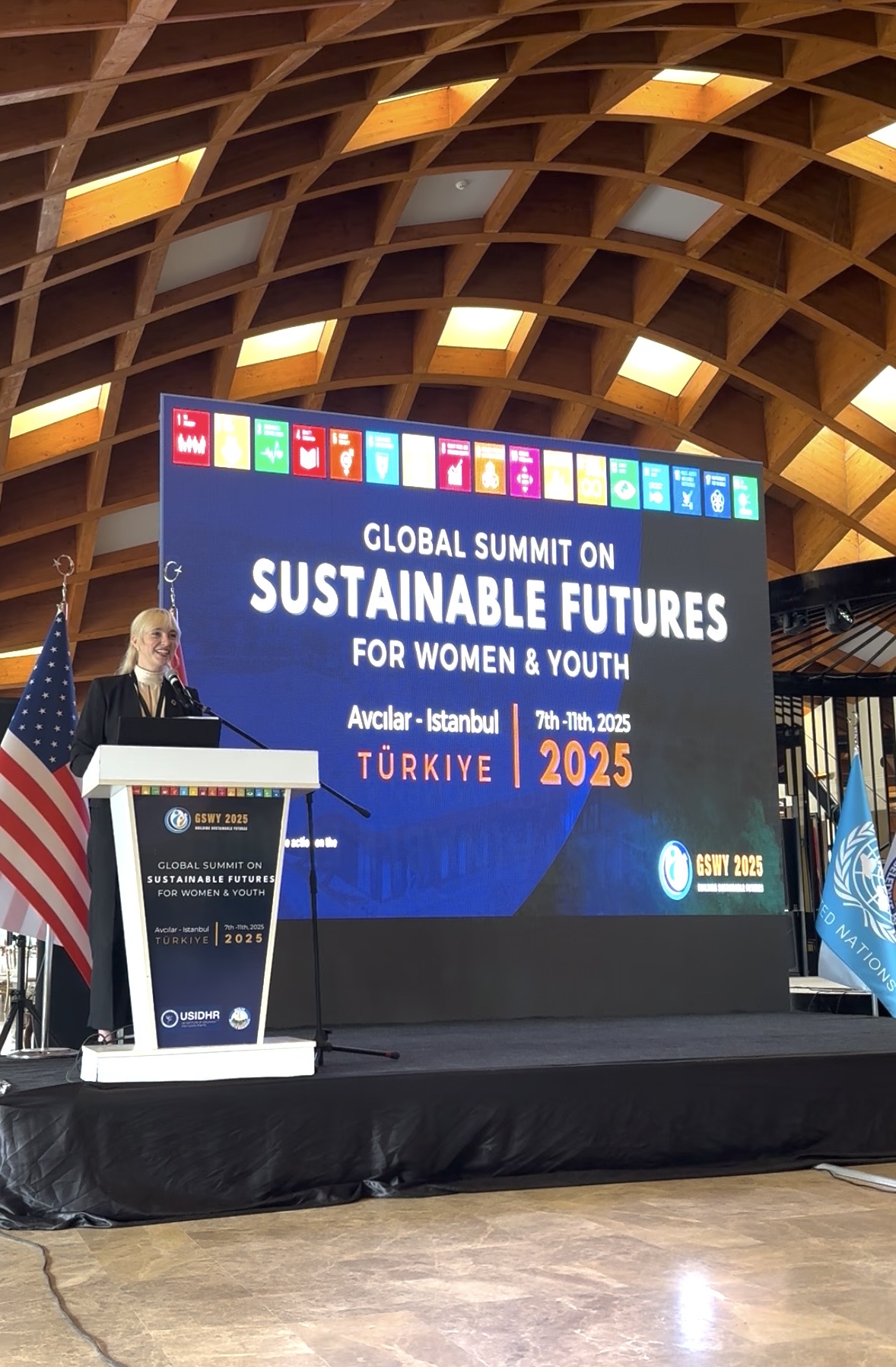 Woman speaking at a podium during a conference with a large screen behind her displaying event details, in a room with a curved wooden ceiling.