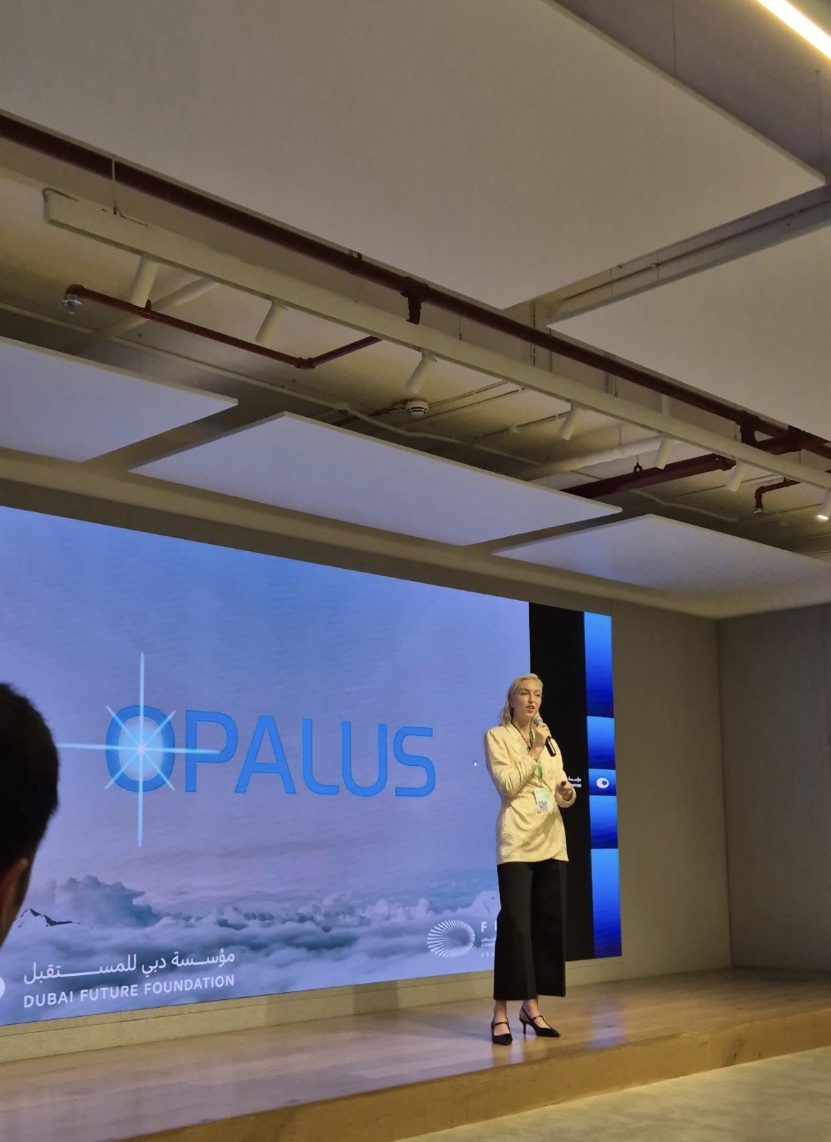 Person standing on stage with a large screen displaying the Opalus logo behind them.