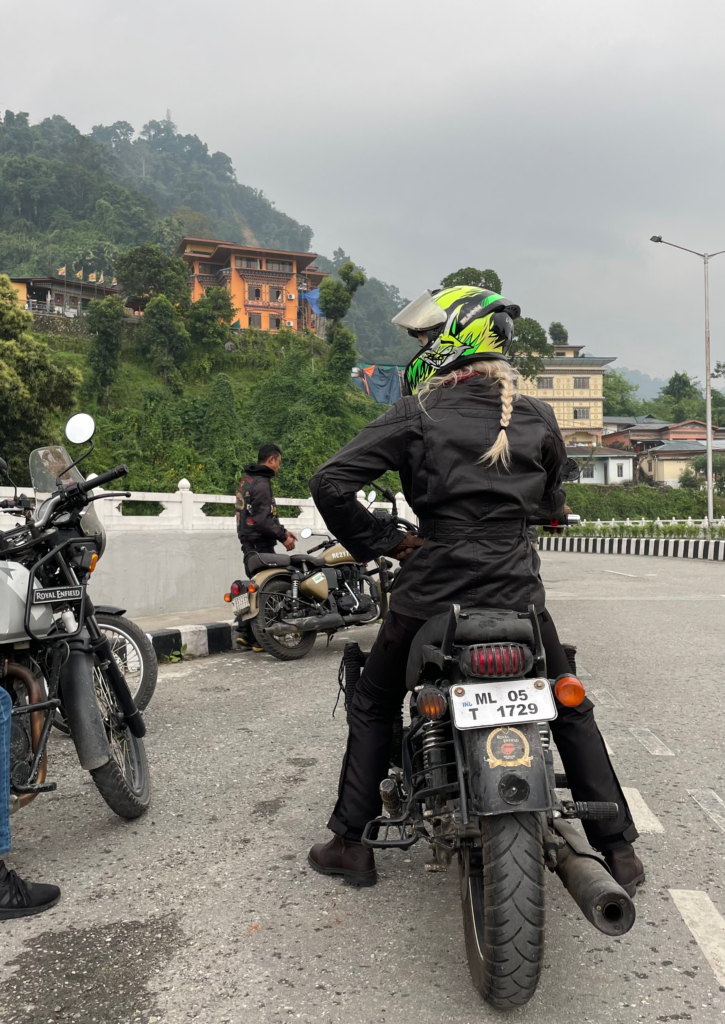 Person wearing a helmet sitting on a motorcycle on the road with other motorcycles and buildings in the background.