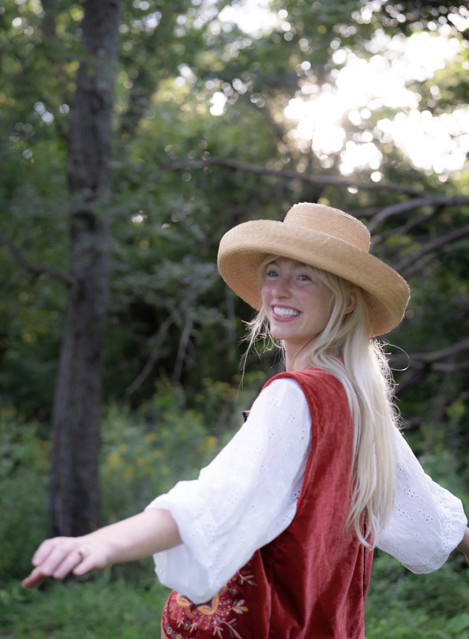 Young woman with long blonde hair wearing a wide-brimmed hat and red vest outdoors, smiling, with trees and sunlight in background.