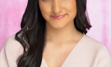 Young woman with long dark hair, smiling, wearing a light pink top, against a pink background.