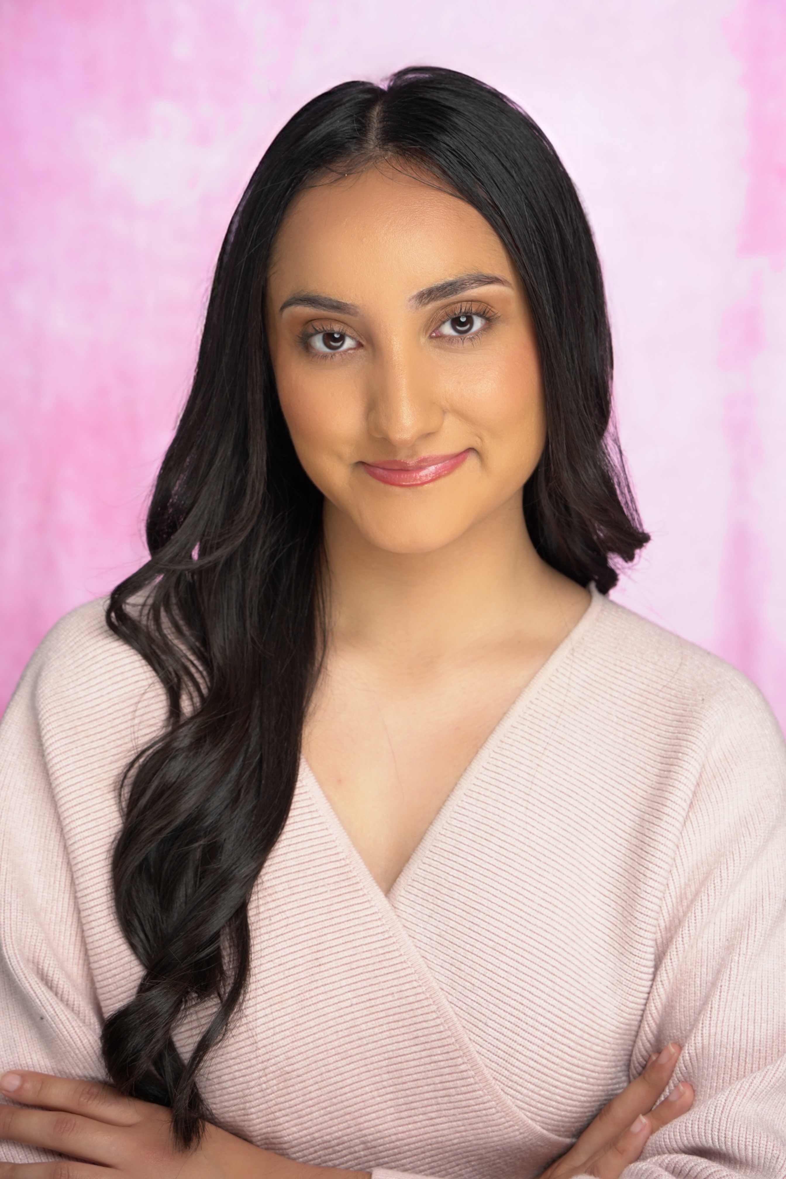 Young woman with long dark hair, smiling, wearing a light pink top, against a pink background.