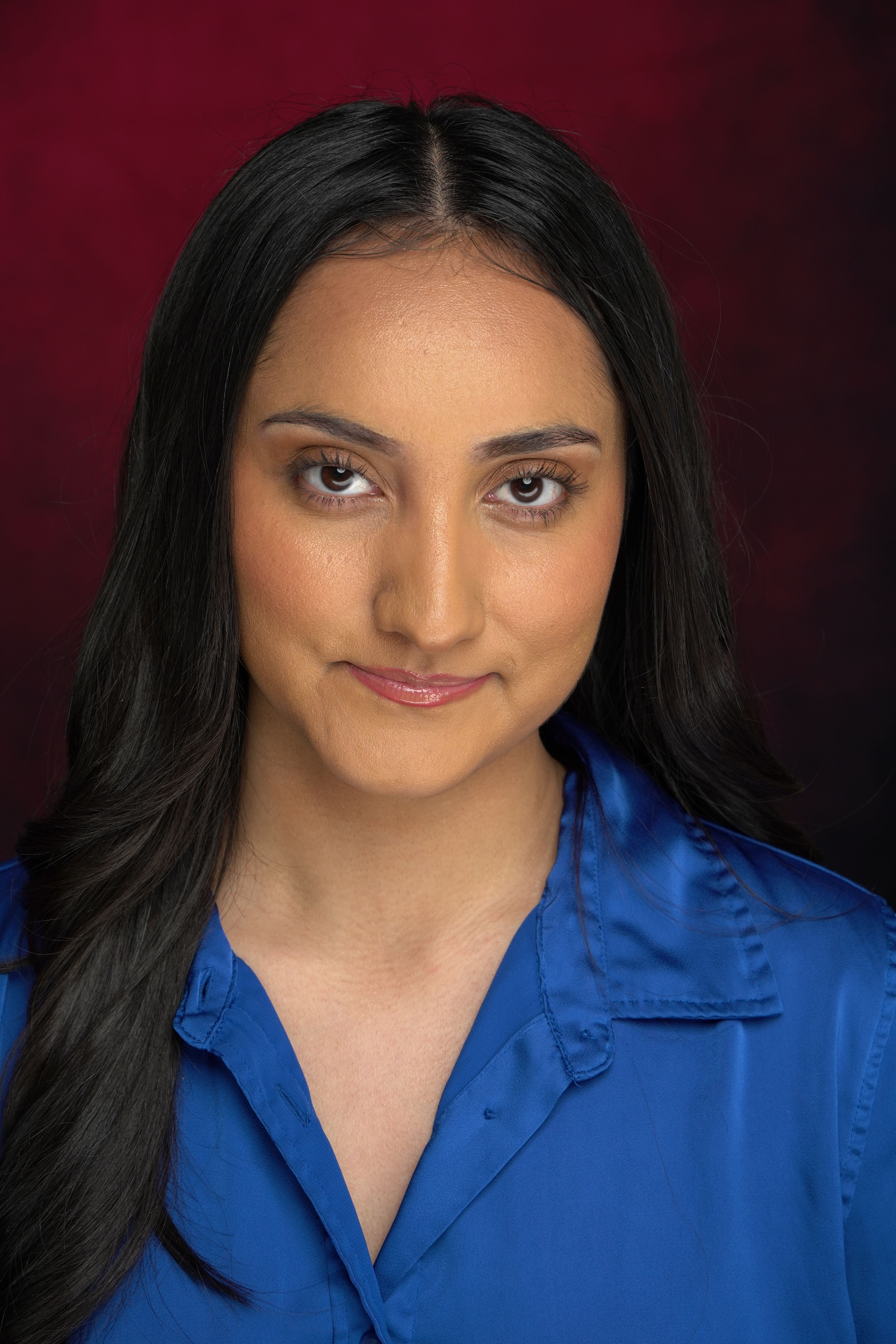 Portrait of a woman with long dark hair wearing a blue top, against a dark red background.