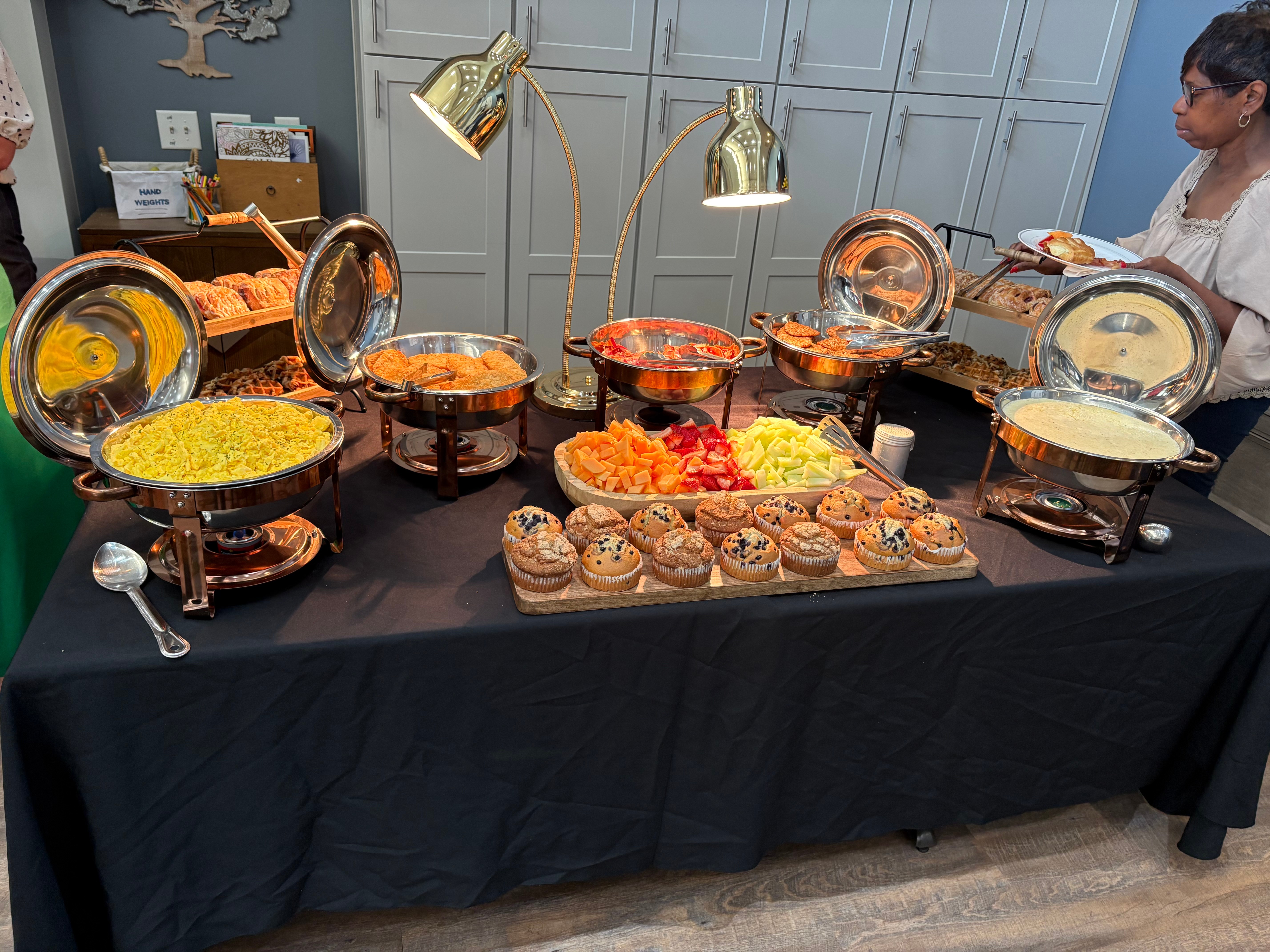 Buffet table with chafing dishes containing rice, vegetables, and desserts, with a woman serving herself in the background.