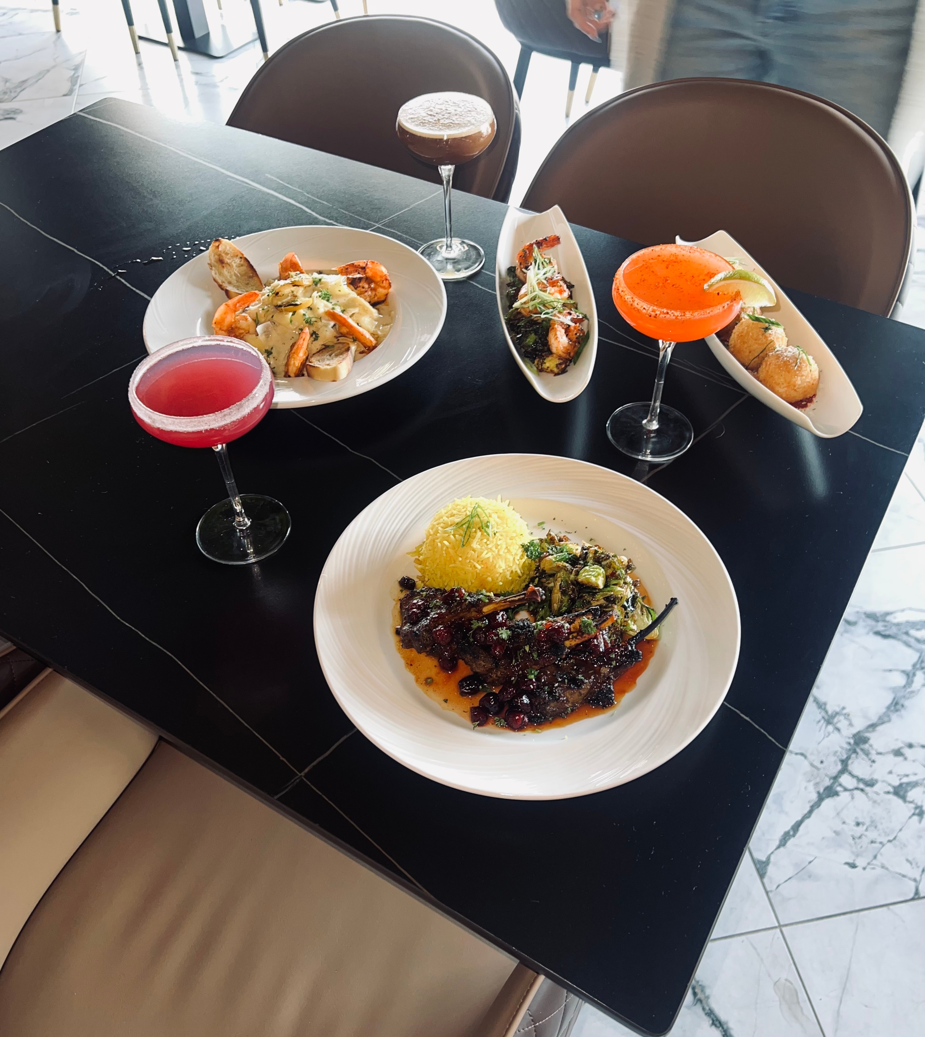 Three plates of food and three colorful drinks on a black table in a restaurant, with chairs and a marble floor visible.