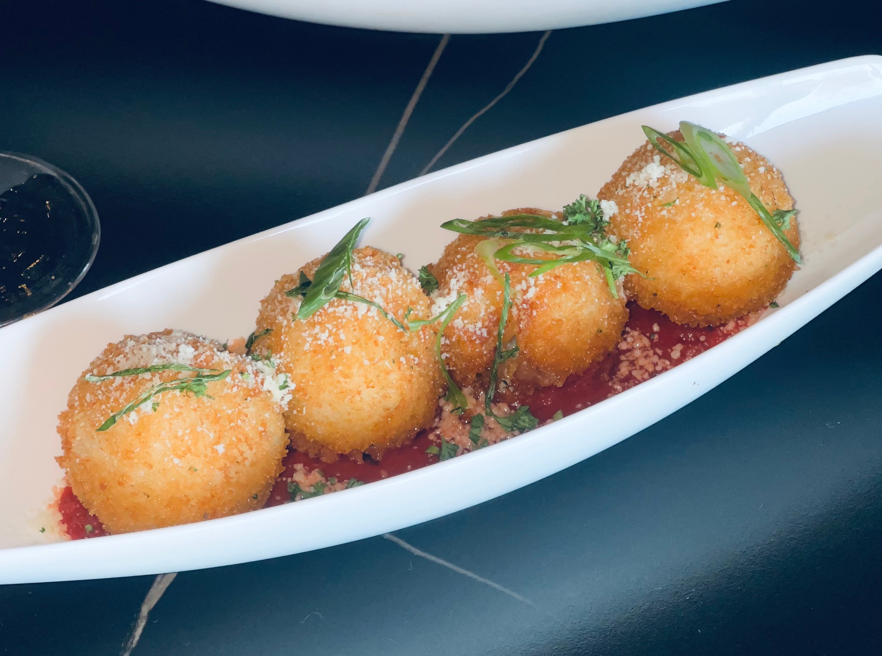 Four breaded, fried balls garnished with green herbs on a white elongated dish, with a dark background.