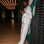 Young man in white tracksuit leaning against a tiled wall, holding a phone, in a modern interior with decorative lighting.