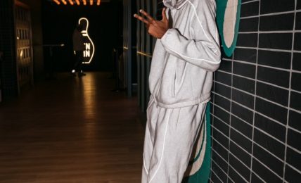 Young man in white tracksuit leaning against a tiled wall, holding a phone, in a modern interior with decorative lighting.