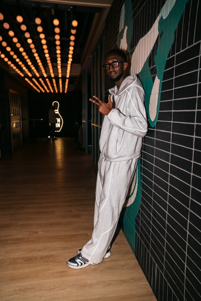 Young man in white tracksuit leaning against a tiled wall, holding a phone, in a modern interior with decorative lighting.