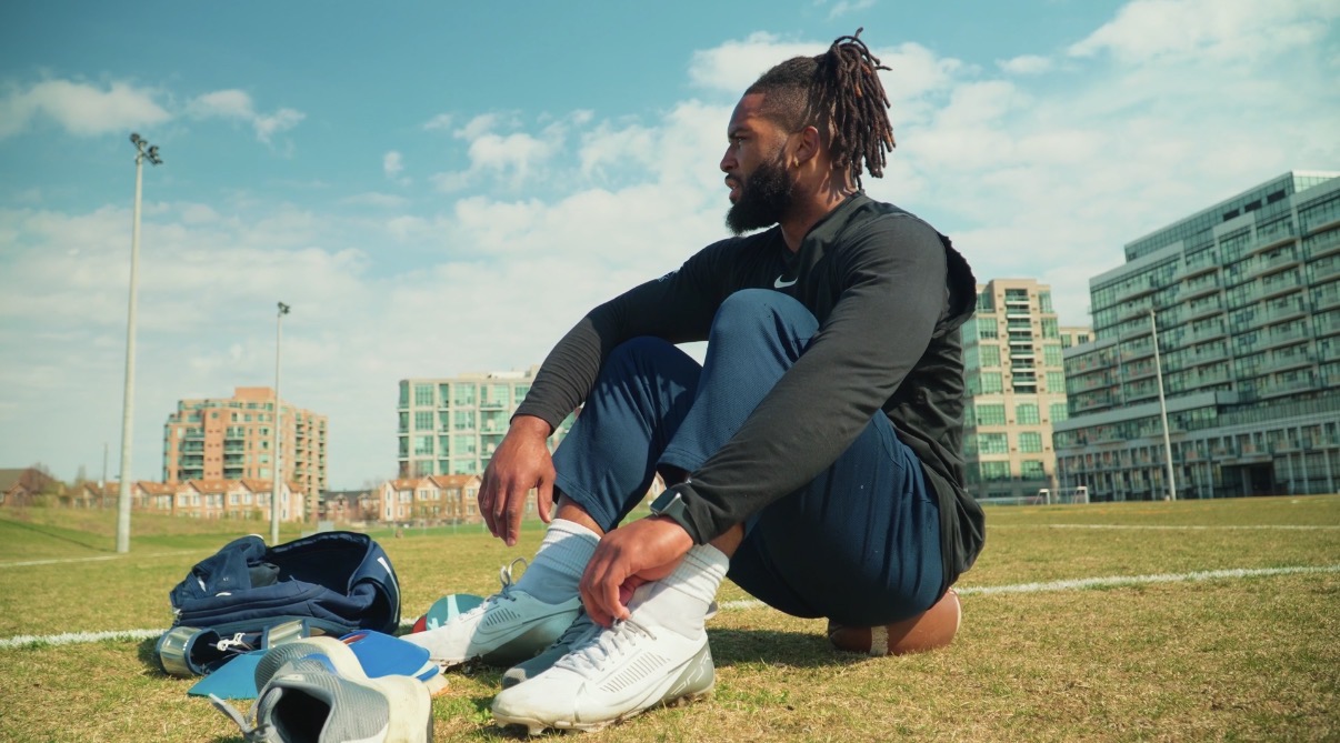 Person sitting on grass tying shoelaces outdoors with buildings and blue sky in background.