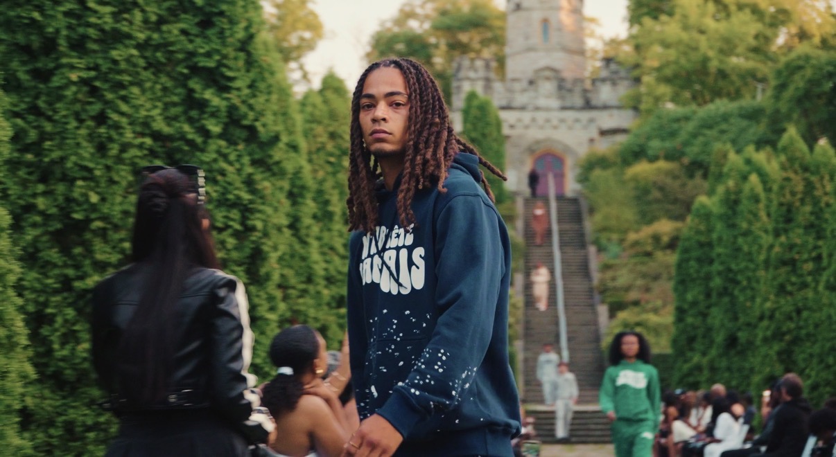 Young woman with dreadlocks in a blue hoodie standing outdoors with people and a castle in the background.