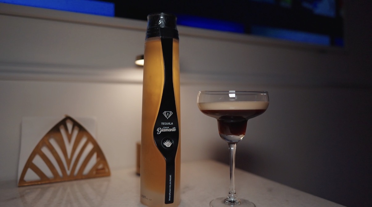 A tall, slim bottle and a glass with dark liquid on a table, with a decorative wooden object in the background.
