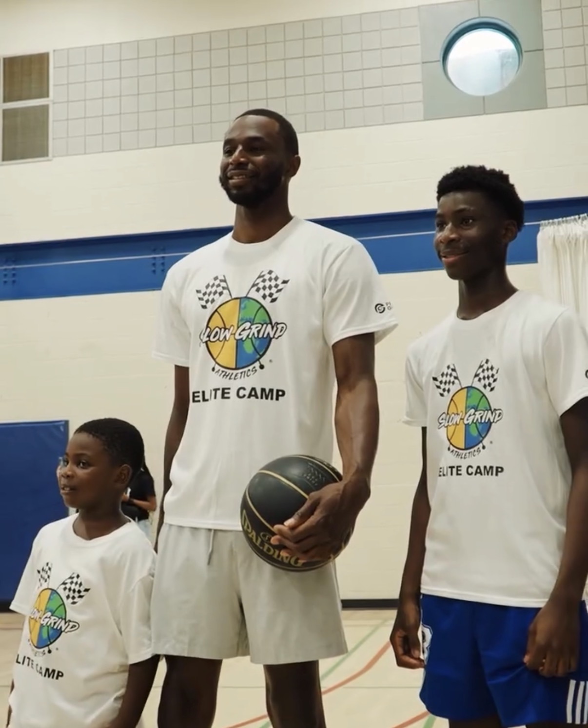 Three people in white T-shirts with a colorful logo, standing indoors, smiling, with a basketball, in a gymnasium.