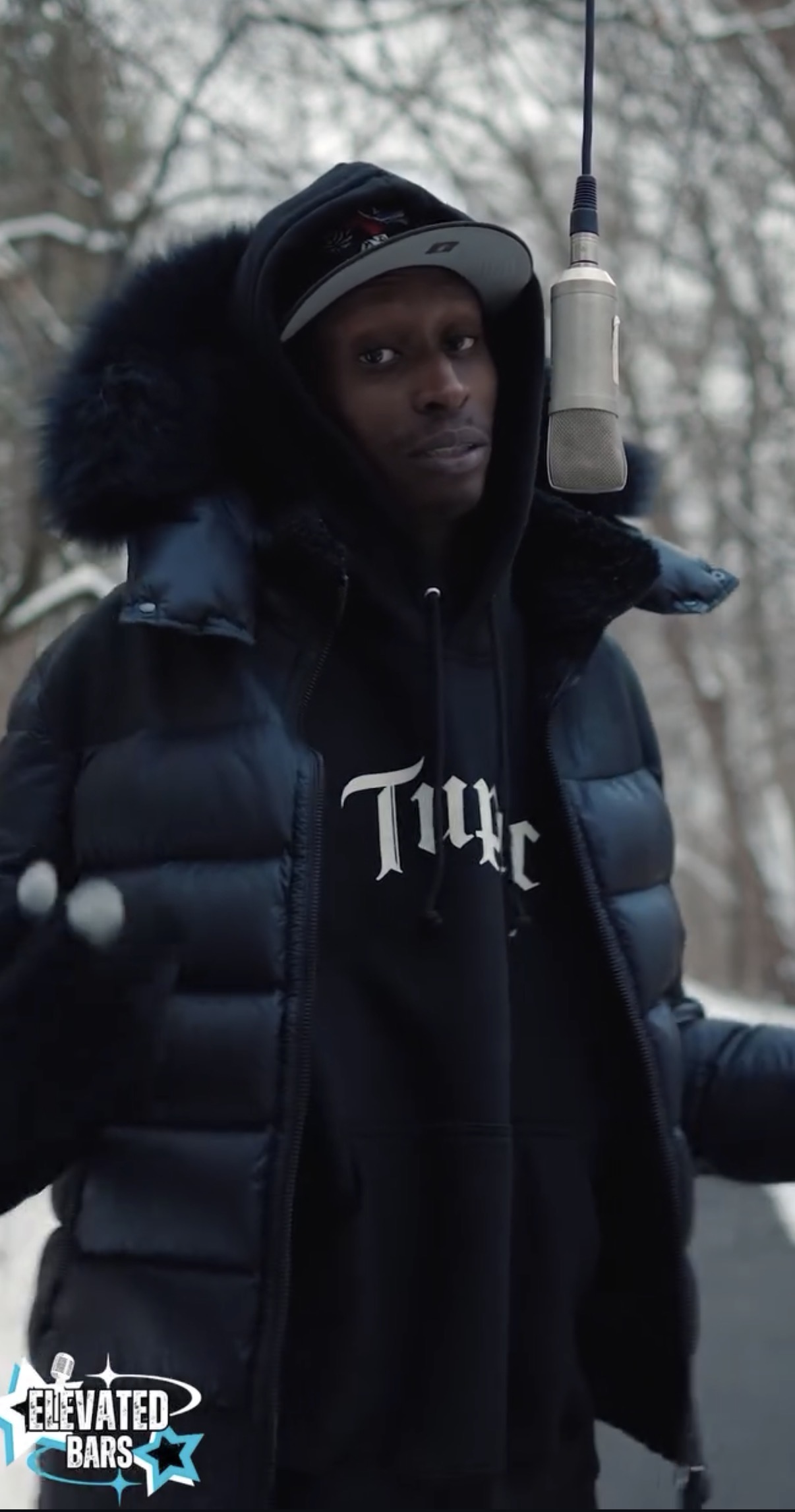 Person wearing a black hoodie and puffer jacket outdoors, with a microphone hanging above, in a winter setting.