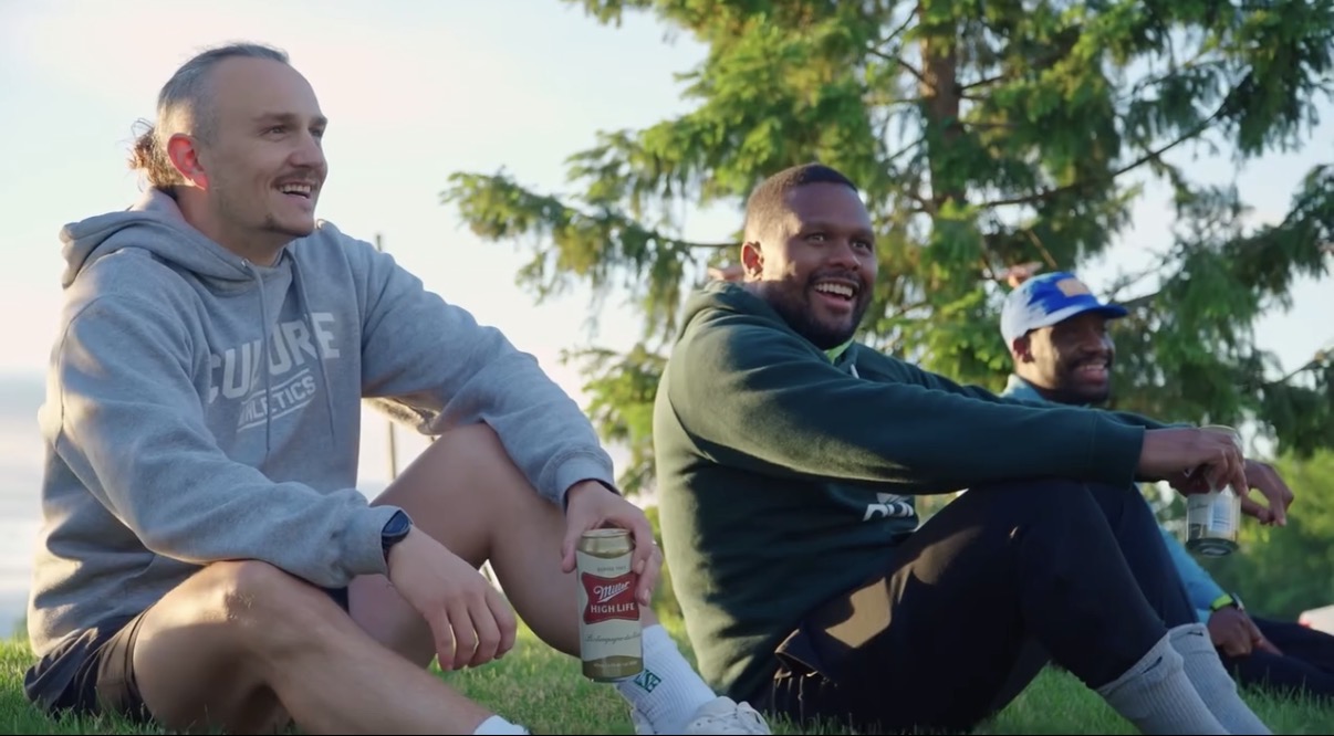Three people sitting outdoors on grass, smiling, with trees in the background.