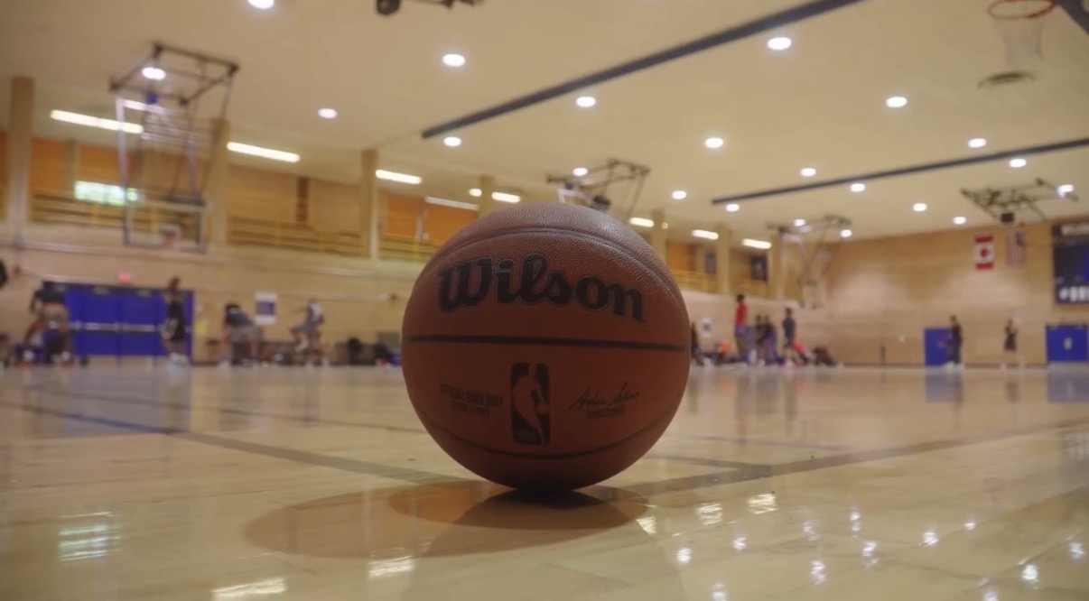 Basketball on a court with players and equipment in the background, indoor gymnasium setting.