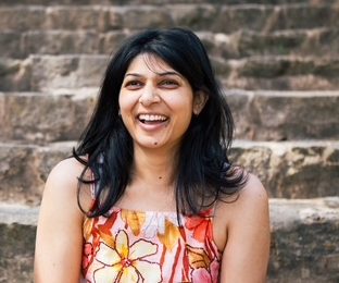 Young woman with dark hair smiling, wearing a floral sleeveless top, sitting on stone steps outdoors.