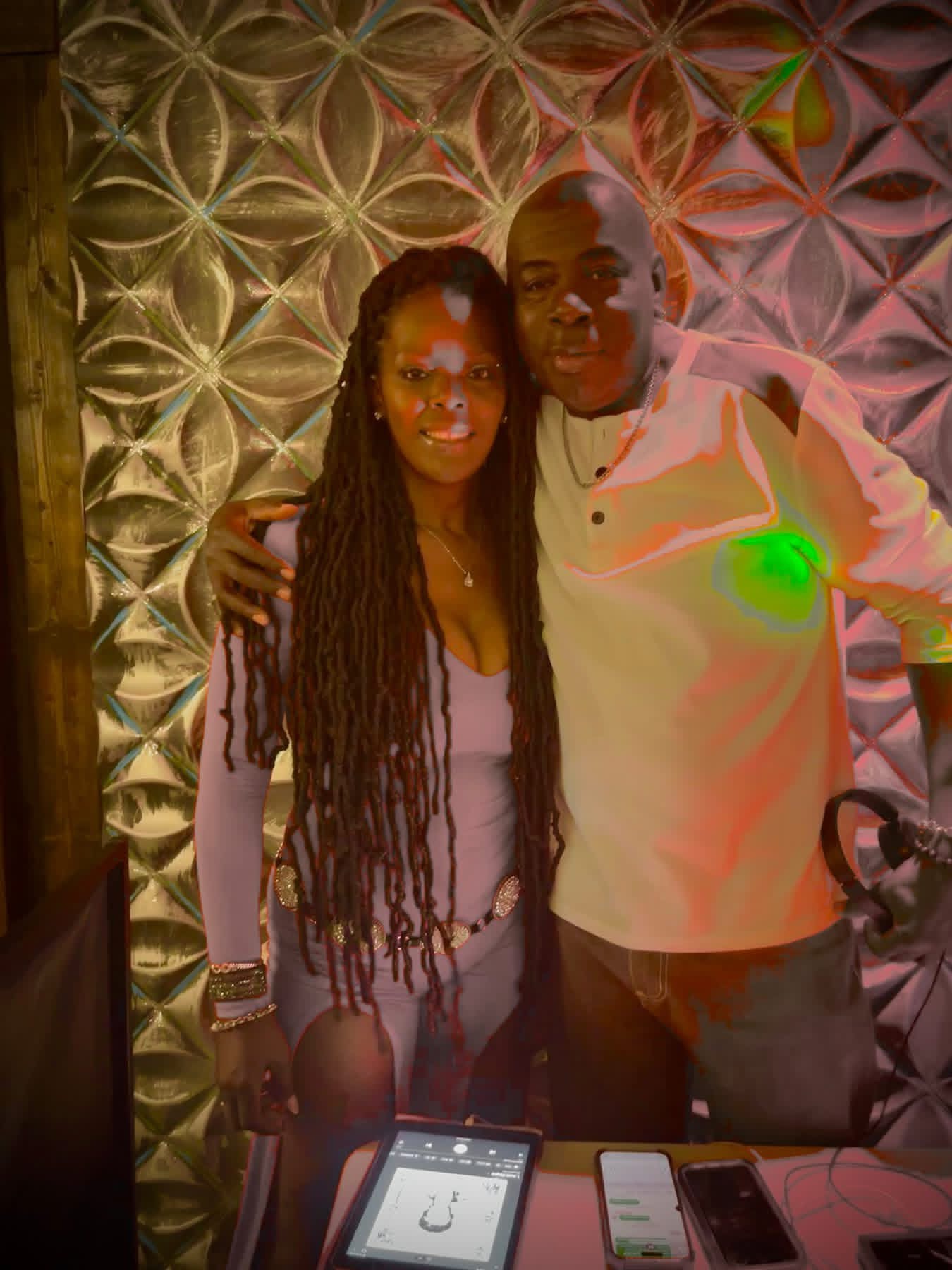 Two people standing close together, smiling, with a patterned wall background, one with long dreadlocks, the other in a white shirt.