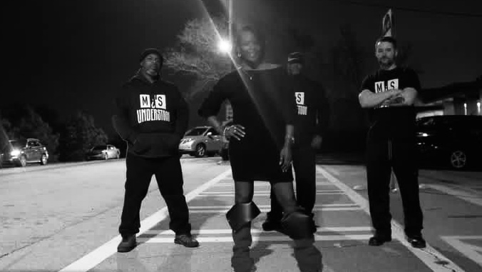 Three people standing in a parking lot at night, wearing dark clothing and hoodies, with cars in the background.
