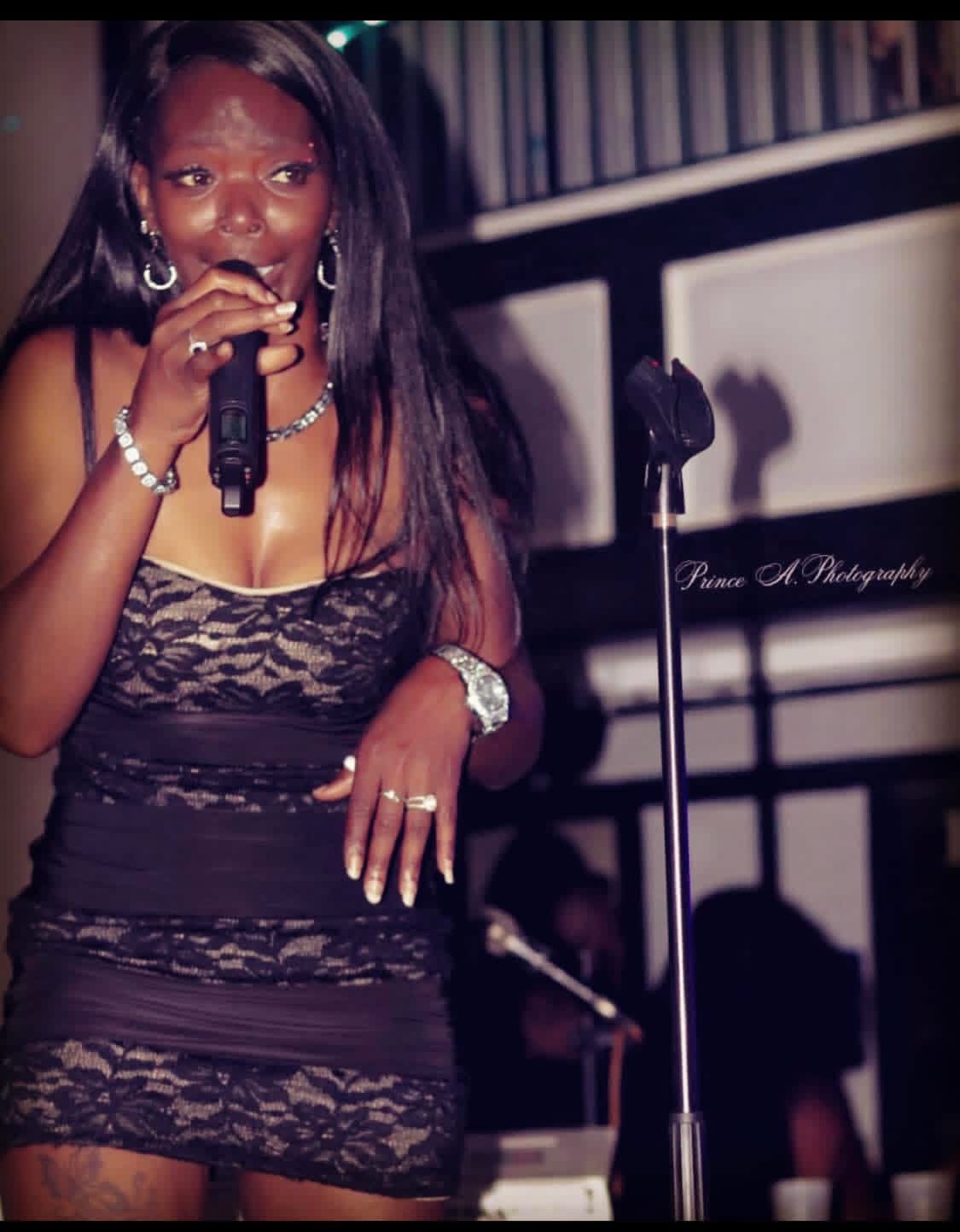 Woman singing into microphone, wearing black lace dress, jewelry, in a dimly lit setting with a microphone stand nearby.