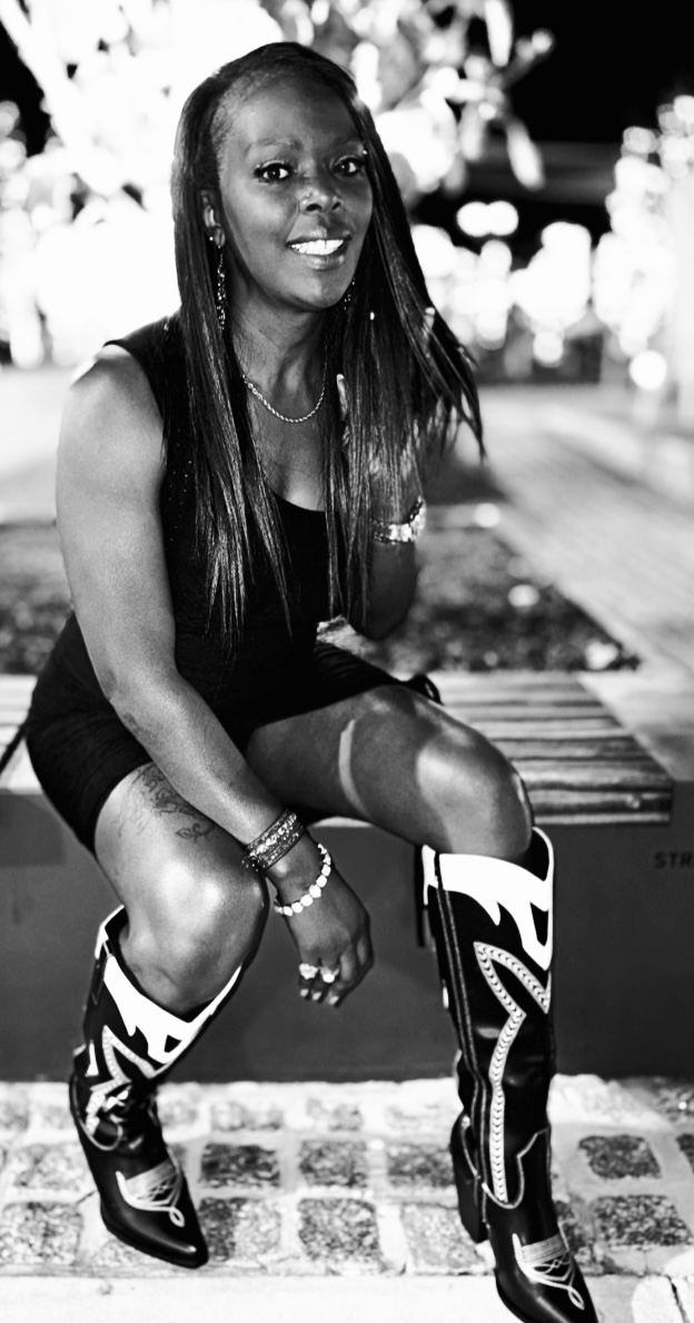 Woman sitting on a bench outdoors, smiling, wearing boots and accessories, in black and white.